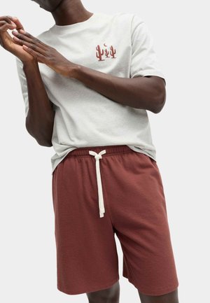 Mann trägt ein hellgraues T-Shirt mit braunen Kaktusmotiven und weinrote Shorts mit Kordelzug, richtet seine Hände.