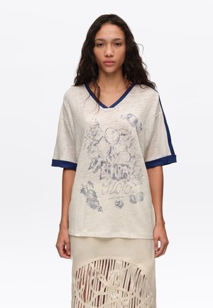 Mujer con cabello oscuro y rizado que lleva una camisa beige sobredimensionada con ribete azul y estampado de frutas, combinada con una falda beige con detalles recortados.