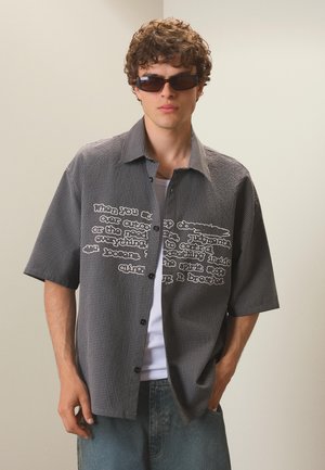 Jonge man met krullend haar die donkere zonnebrillen draagt, een kortemouwen geruit hemd met witte tekst, wit ondershirt en blauwe spijkerbroek, ontspannen staand.