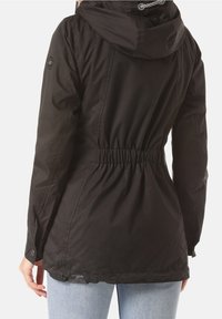 Kvinna som bär en åtsittande svart huva-jacka med elastisk midja och långa ärmar, i kombination med ljusblå jeans, visad bakifrån.