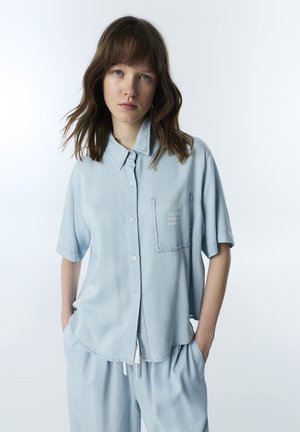 Jeune femme aux cheveux châtain mi-longs portant une chemise ample bleu clair à manches courtes et un pantalon assorti, les mains dans les poches.