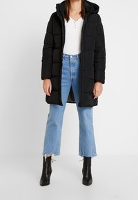 Abrigo acolchado negro con capucha, con texturizado acolchado y cremallera frontal. Llevado sobre una camiseta blanca y unos jeans de campana azul claro, combinado con botas negras.