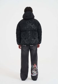 Svart puffjacka med huva, med en stor grafisk design på baksidan. Grå vida jeans med färgglad broderad detalj på sidan.