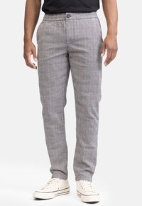 Mann in grauen karierten, eng anliegenden Hosen und weißen Canvas-Sneakern, steht mit entspannten Händen an den Seiten.