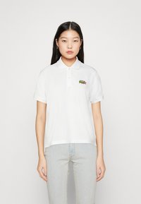 Camisa polo blanca con cuello clásico, mangas cortas y un logo bordado en verde en el pecho. Material de algodón suave, corte relajado.