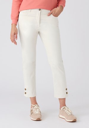 Donna che indossa pantaloni a taglio corto color avorio con bottoni tartarugati sull'orlo, una maglietta a maniche lunghe color corallo e scarpe da ginnastica beige con stringhe.