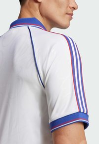 Biele športové tričko s modrým golierom, červenými a modrými pruhmi na rukávoch, s jemnými vzormi na textúre látky.