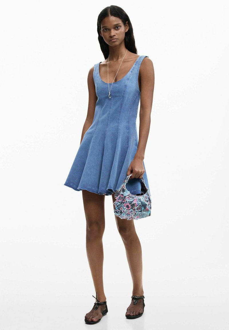 Woman wearing sleeveless blue denim dress, holding floral-patterned handbag, and wearing black mesh shoes and a silver necklace.