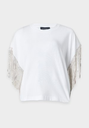 Mujer de pie con una camiseta blanca de manga corta con flecos de cadena y pantalones negros de pierna ancha con zapatos metálicos puntiagudos.