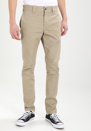 Personne portant un pantalon beige slim fit et des baskets en toile noires et blanches, debout devant un fond clair uni.