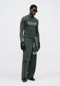 Graues Langarmshirt mit dem Text "DECONSTRUCTION", kombiniert mit einem drapierten grauen Rock über grauen Hosen, Lederhandschuhen und dunklen Sonnenbrillen.
