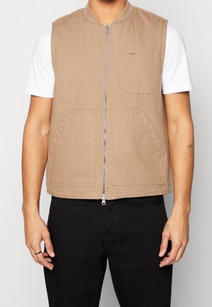 Mann trägt beige ärmellose Weste mit Reißverschluss über weißem kurzärmligem Hemd und schwarzen Hosen, steht vor schlichten Hintergrund.