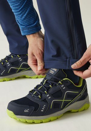 Persona ajustando el puño de unos pantalones azul marino para exteriores sobre zapatos de senderismo impermeables negros y neón verdes sobre una superficie clara.