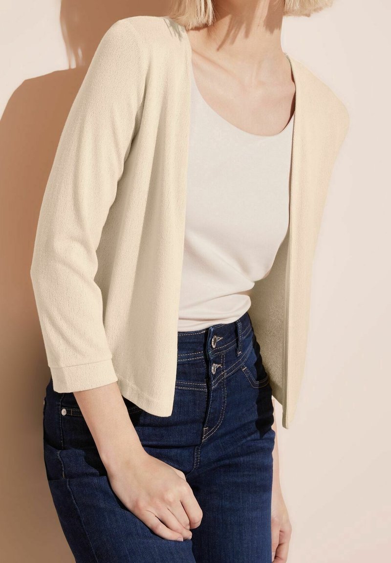 Lichtbeige openvallend vest met driekwart mouwen, gestructureerde stof, gecombineerd met een witte top en donkerblauwe high-waisted jeans.