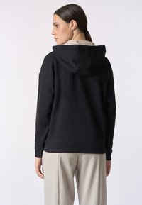 Felpa nera con una texture morbida, dotata di cappuccio regolabile con coulisse e polsini a coste, abbinata a pantaloni chiari per creare contrasto.