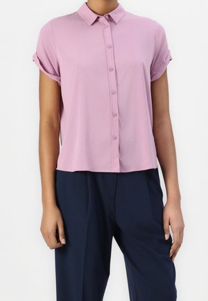 Femme portant un chemisier rose à manches courtes avec des poignets retroussés et des boutons, ainsi qu'un pantalon large bleu marine, debout devant un fond blanc.