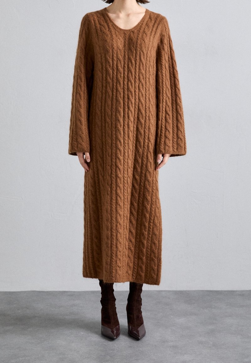 Personne portant une longue robe-pull en maille torsadée marron avec de larges manches et des bottes à talons hauts pointues marron foncé, debout sur un sol gris.