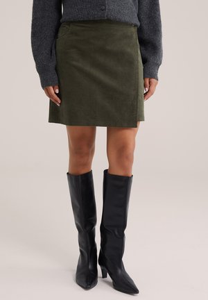Persona con una falda de pana verde oscuro, suéter gris de punto y botas negras de tacón hasta la rodilla, de pie frente a un fondo liso.