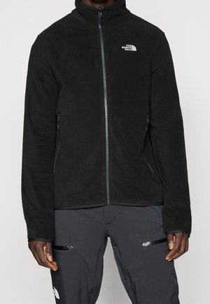 Czarna bluza polarowa na pełny zamek błyskawiczny z logo The North Face na piersi, noszona z ciemnymi spodniami z kieszeniami na udach zapinanymi na zamek.