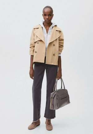 Beige double-breasted trench coat with wide collar, gray hoodie underneath, dark gray pants, and brown shoes; holding a gray handbag.