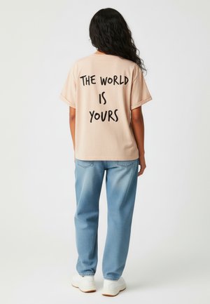 lichtroze oversized t-shirt met zwarte tekst "DE WERELD IS VAN JOU" op de achterkant, gedragen met lichtblauwe denim broek en witte sneakers.