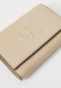 Portefeuille en cuir beige avec logo "CK" embossé, design pliable et bords cousus. Texture lisse avec une esthétique minimaliste.