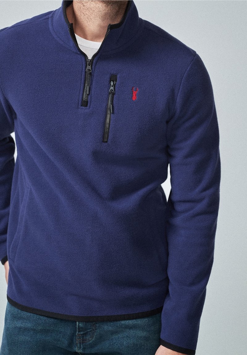 Marineblauer Fleece-Pullover mit einem halben Reißverschlusskragen, ausgestattet mit einem kleinen roten Logo und schwarzen Reißverschlussdetails. Glatte Textur und taillierte Passform.