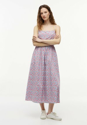 Jonge vrouw met krullend haar die een mouwloze, bloemen midi-jurk en witte sneakers draagt, met gekruiste armen op een effen achtergrond.