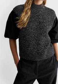 Šedý pletený svetřík bez rukávů s vysokým límcem, s texturovanou látkou a kontrastními černými krátkými rukávy. Nošený přes tmavé na míru šité kalhoty.