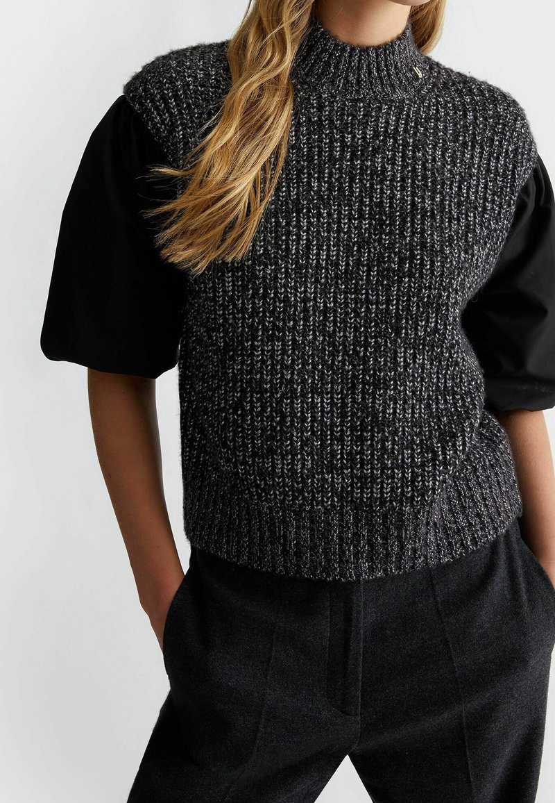 Šedý pletený svetřík bez rukávů s vysokým límcem, s texturovanou látkou a kontrastními černými krátkými rukávy. Nošený přes tmavé na míru šité kalhoty.
