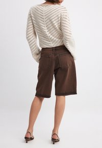 Suéter de punto abierto en beige claro con un patrón de chevron, combinado con shorts de mezclilla marrones de corte relajado y dos bolsillos traseros. Tacones negros.