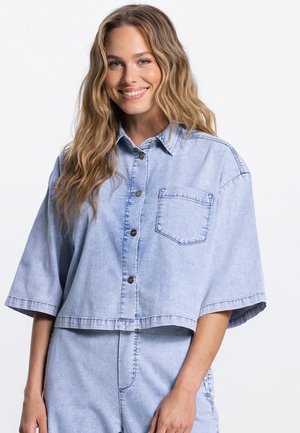 Lachende vrouw met lang golvend haar, gekleed in een lichtblauw oversized denim overhemd met knopen aan de voorkant en bijpassende denim broek.