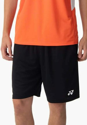 Person trägt schwarze Sportshorts mit weißem Logo am linken Bein und ein oranges Sportshirt, steht mit entspannten Händen.