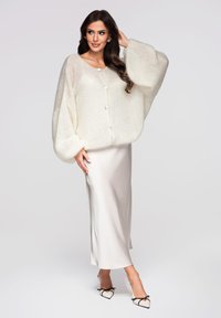 Cardigan oversized blanc en maille avec de gros boutons, porté par-dessus une jupe en satin crème, dotée d'une texture lisse et de manches ballon.