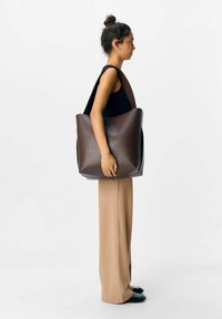 Femme avec un chignon foncé portant un haut sans manches noir et un pantalon beige se tient de profil, portant un grand sac fourre-tout en cuir marron sur l'épaule.