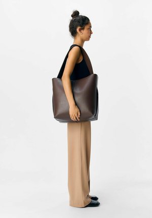 Femme avec un chignon foncé portant un haut sans manches noir et un pantalon beige se tient de profil, portant un grand sac fourre-tout en cuir marron sur l'épaule.