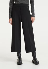 Pantalon noir à jambes larges en tissu lisse, avec une taille haute et des lignes épurées, associé à des bottines noires.