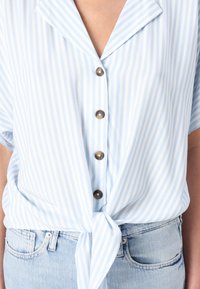 Light blue and white striped shirt with short sleeves, a lapel collar, five metallic buttons, and a tied front detail over denim jeans.
