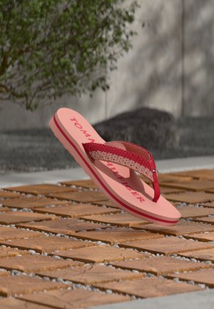 Infradito rosa Tommy Hilfiger con cinturini intrecciati rossi e beige sospesi sopra un pavimento in legno piastrellato all'aperto.