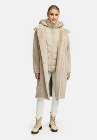 Lange, beige nepbontjas die over een licht gewatteerde jas met capuchon ligt. Gecombineerd met witte broeken en beige veterschoenen.