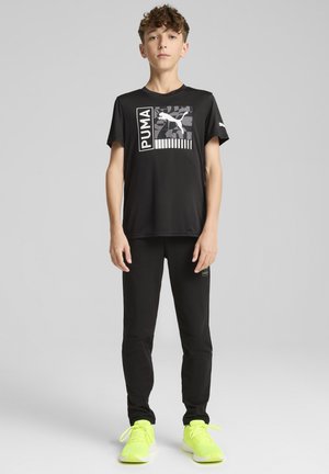 Camiseta atlética negra con un logotipo gráfico de Puma y un diseño estampado, combinada con pantalones negros y zapatillas amarillas brillantes.