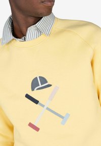 Sudadera amarilla con cuello redondo, que presenta un bordado de una gorra gris y mazos cruzados en rosa, azul y negro. Camisa de rayas debajo.