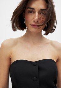 Mustat olkaimettomat topit, joissa on teksturoitu kangas, keskellä kaksi pyöreää nappia, korostaen istuvaa muotoa ja sulavaa kaula-aukkoa.