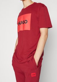 Camiseta de algodón color burdeos con un logotipo rectangular rojo que lleva "HUGO" en negro. Manga corta y corte estándar. Textura suave.