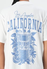 Balta marškiniai su mėlynu grafiniu atvaizdu, vaizduojančiu gėrimo puodelį, citrusinius vaisius ir užrašą "Los Angeles CALIFORNIA" ryškiu šriftu.