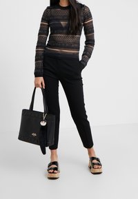 Svart genomskinlig långärmad topp med geometriska mönster, svarta smalna byxor, svart tote-väska med charm, och plattformssandaler med spännen.