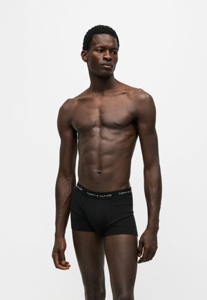 Modelo masculino de piel oscura con calzoncillos bóxer negros Tommy Hilfiger, de pie sobre un fondo blanco liso.