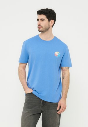 Άνδρας με κοντά σκούρα μαλλιά, φοράει μπλε μπλουζάκι με ένα μικρό πολύχρωμο σχέδιο κυμάτων στο αριστερό στήθος, στέκεται με το ένα χέρι στην τσέπη.