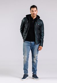 Černá kožená bunda s kapucí, modré roztrhané džíny a tmavé tenisky. Bunda má zipové kapsy a přiléhavý střih.