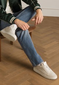 Beige Wildledersneaker mit einer strukturierten Oberfläche, weißer Gummisohle und minimalistischem Design, kombiniert mit hellblauen Jeans.